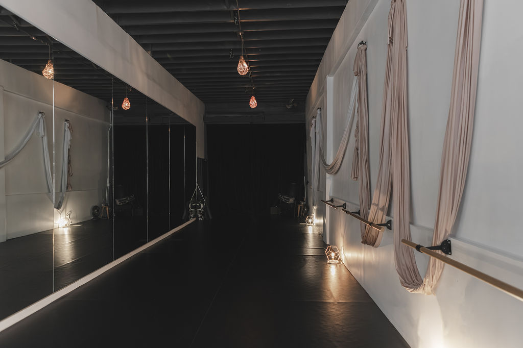 Empty aerial yoga studio with black padded floors, a mirrored wall on the left, and beige aerial hammocks draped along a white wall with mounted ballet barres. Warm pendant lights hang from an exposed dark ceiling, and heavy black curtains cover the back wall.