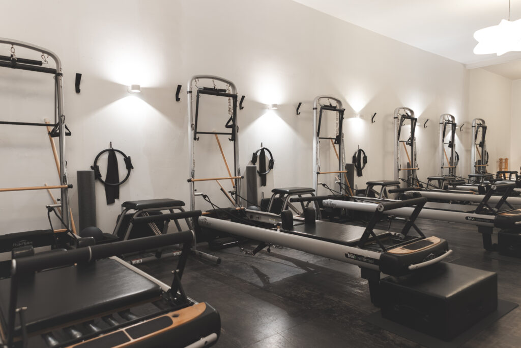 Bright Pilates studio with multiple reformers arranged in rows on dark wood floors, each paired with a black box. Tower units with springs and bars are mounted along a white wall, with neatly hung straps and rings beneath soft wall lighting.
